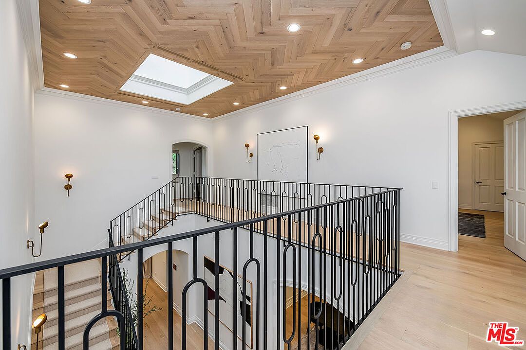 This interior shot showcases a well-lit hallway and staircase with a modern aesthetic. The ceiling features a unique herringbone wood pattern with a skylight, complemented by white walls and black metal railings. The staircase is carpeted, and the hallway leads to various rooms, creating a bright and inviting transition space.