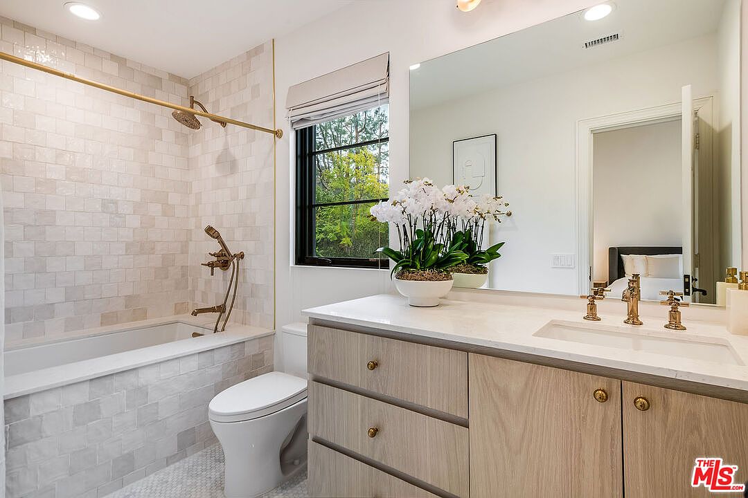 This is a bright and stylish bathroom featuring a bathtub with a shower, a toilet, and a vanity with a sink. The walls are tiled in a neutral color, and the fixtures are gold. A window provides natural light, and a mirror reflects the space, creating an open and airy feel.