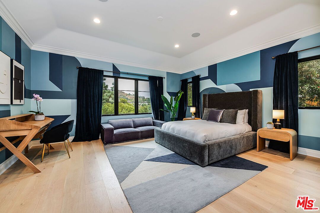 This is a primary bedroom featuring a large bed with a high headboard, a gray sofa, and a wooden desk. The walls are painted in a geometric pattern of blues and the room is well-lit with natural light from the windows. The overall style is modern and sophisticated.