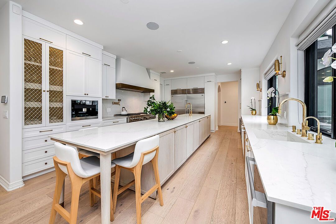 This is a bright and spacious kitchen featuring white cabinetry, a large center island with a white countertop and seating, and stainless steel appliances. The kitchen has hardwood floors and gold fixtures, creating a luxurious and modern feel. The perspective is from a wide angle, showcasing the length of the kitchen and the natural light coming in from the window.