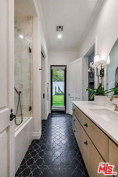 This is a bathroom featuring a modern design with a black and white patterned tile floor. The vanity has light wood cabinets and a white countertop with a sink. A shower with glass doors is on the left, and a door leading to the outside is at the end of the hallway, bringing natural light into the space.