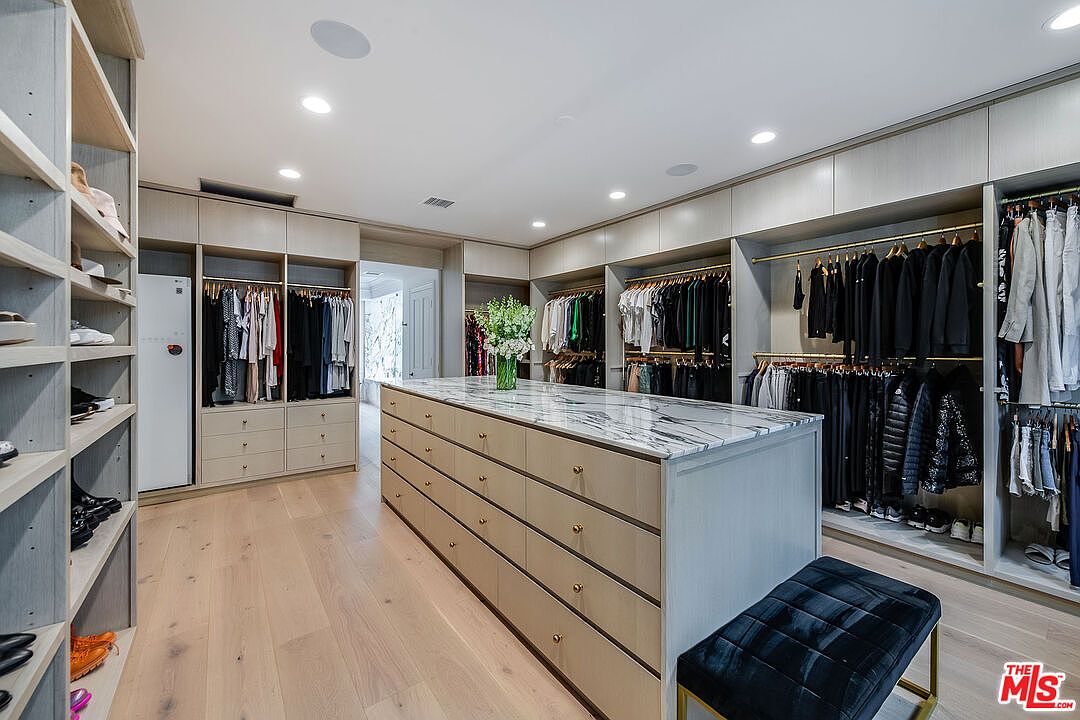 This is a spacious walk-in closet featuring custom cabinetry with drawers and shelving, providing ample storage for clothing and accessories. A large center island with a marble countertop offers additional surface space, while a plush bench provides a comfortable seating area. The overall design is luxurious and well-organized, creating an elegant dressing space.