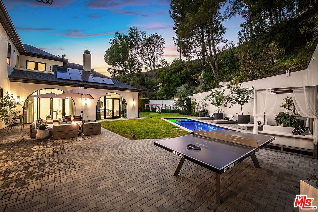 This image showcases a luxurious backyard featuring a modern home with arched windows and solar panels. The outdoor space includes a well-manicured lawn, a swimming pool, a lounge area with a canopy, and a ping pong table on a brick patio. The scene is set against a backdrop of lush greenery, creating a serene and upscale ambiance.