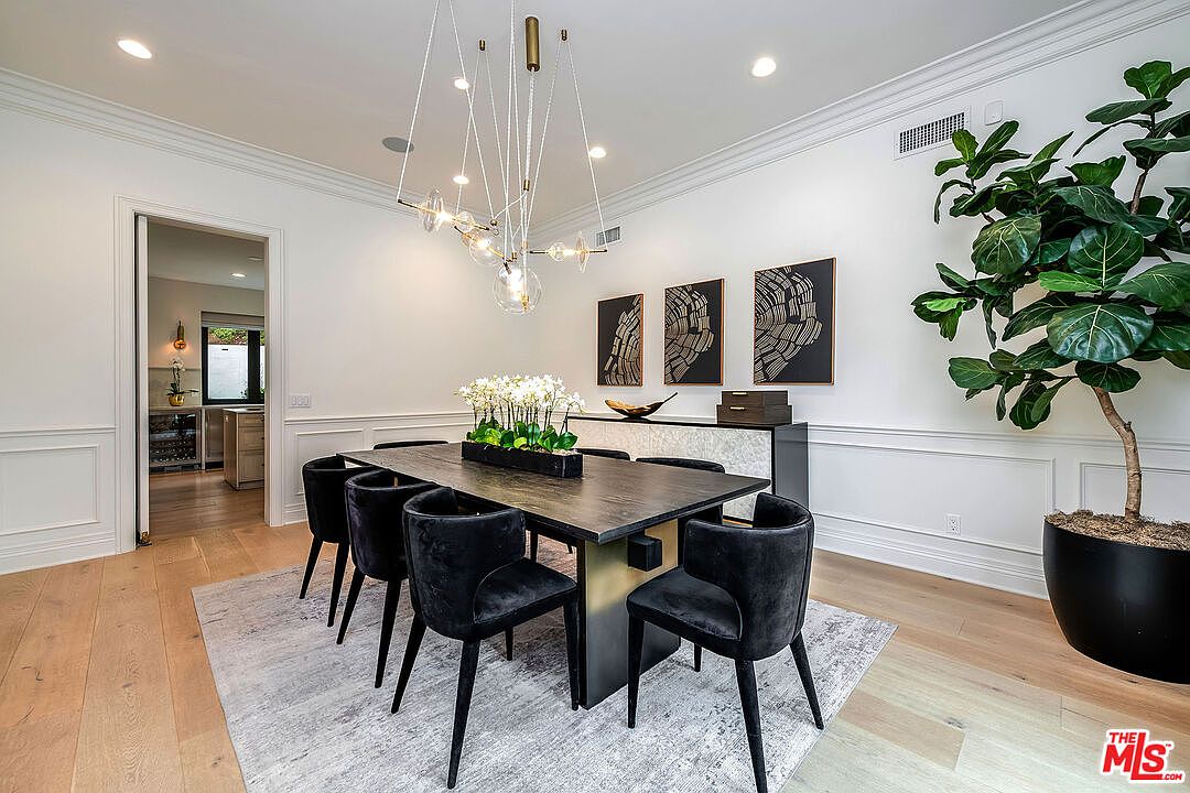 This is an interior shot of a dining room featuring a dark wood table with black velvet chairs, set on a light gray rug. A modern chandelier hangs above the table, and artwork adorns the wall behind a sideboard. A large potted fiddle-leaf fig tree adds a touch of greenery to the space, creating a sophisticated and inviting atmosphere.