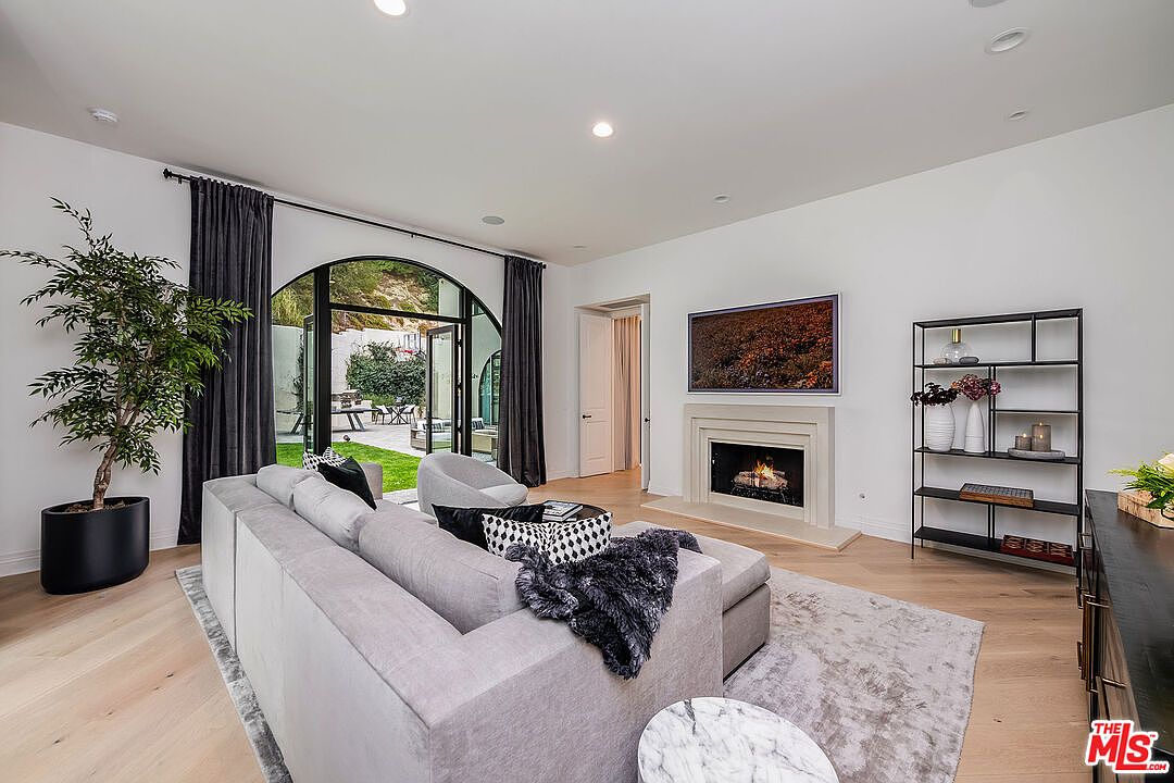 This is an interior shot of a living room featuring a large gray sectional sofa, a fireplace with a television mounted above it, and a black shelving unit. The room has hardwood floors, white walls, and a large arched window/door leading to an outdoor area. The style is modern and minimalist, creating a bright and inviting space.