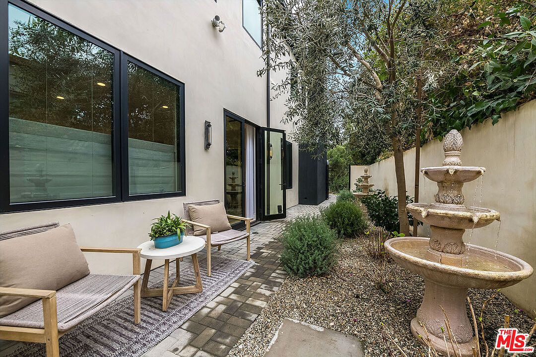 This is a charming exterior shot of a yard or garden area, featuring a stone fountain as a focal point. The space includes seating with chairs and a small table, creating a cozy outdoor living area. The landscaping is well-maintained with various plants and trees, and a pathway leads through the garden, enhancing the overall appeal of the property.