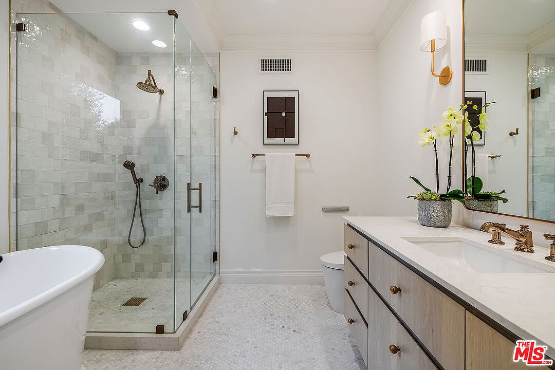 This is a well-lit primary bathroom featuring a glass-enclosed shower with bronze fixtures and tiled walls. A freestanding bathtub is partially visible on the left. The vanity has a light wood finish with bronze hardware and a white countertop, complemented by a large mirror and decorative lighting. The floor is tiled with small hexagonal tiles, adding a touch of elegance.