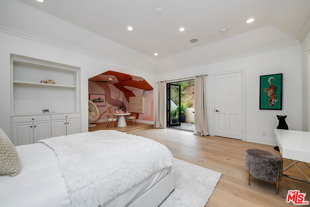 This primary bedroom features a bright and airy atmosphere with white walls and light wood flooring. A cozy bed with a plush white blanket is the focal point, complemented by a built-in shelving unit and an adjacent sitting area with unique wall art. French doors lead to an outdoor space, enhancing the room's appeal.