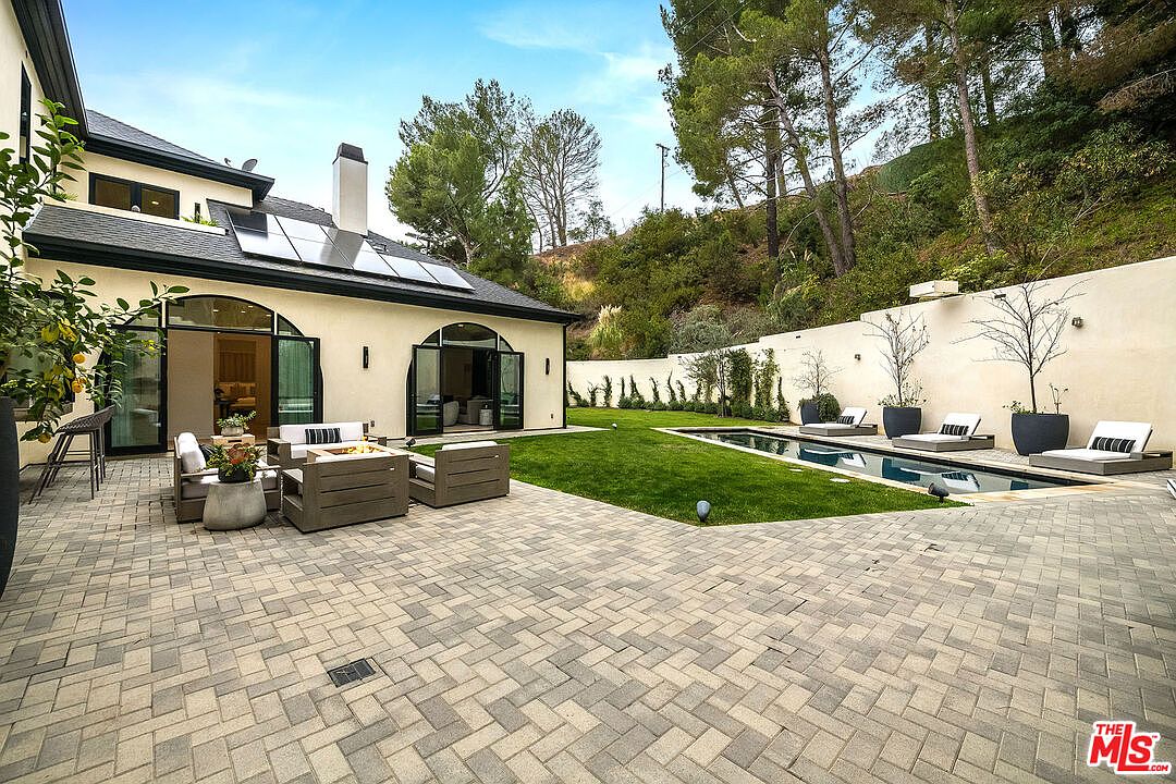 This image showcases a luxurious backyard featuring a modern pool and spa area. The pool is surrounded by a well-manicured lawn and a paved patio with stylish outdoor furniture, including lounge chairs and a seating area with a fire pit. The property is enclosed by a high wall, providing privacy, and the architecture of the house is visible in the background, featuring a contemporary design with large windows and solar panels.
