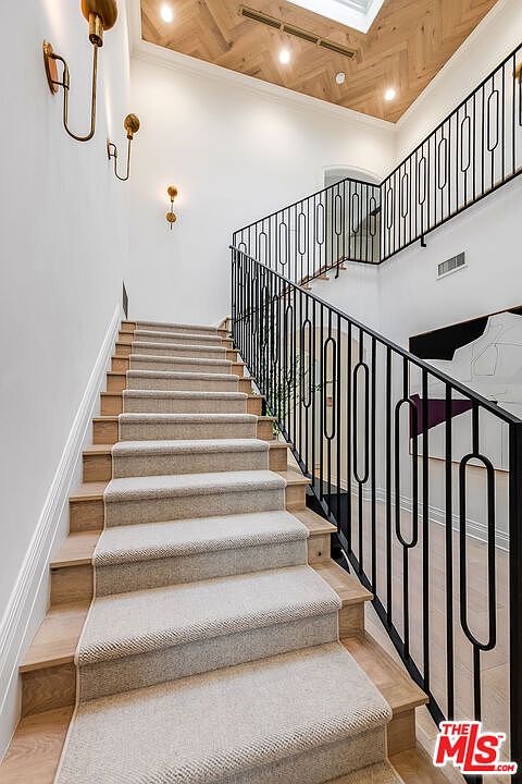 This image showcases a well-lit staircase with carpeted steps and wooden accents. A modern black railing with a unique design adds a touch of elegance. The walls are painted in a neutral tone, complemented by stylish wall sconces, creating a sophisticated and inviting atmosphere.