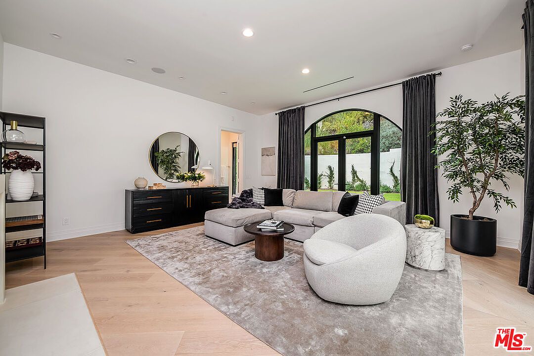 This is a bright and airy living room featuring a large sectional sofa, a round accent chair, and a dark wood console table with a round mirror above it. The room is decorated in neutral tones with pops of black and gold, and a large arched window provides natural light and a view of the outdoors. The overall impression is modern and sophisticated.
