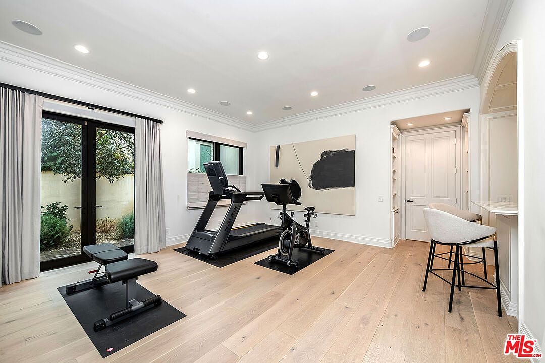This is a well-lit home gym featuring light wood flooring, white walls, and crown molding. The room includes a treadmill, stationary bike, and weight bench, all arranged on black mats. A large abstract painting hangs on the wall, and a glass door leads to an outdoor area, creating a bright and inviting workout space.