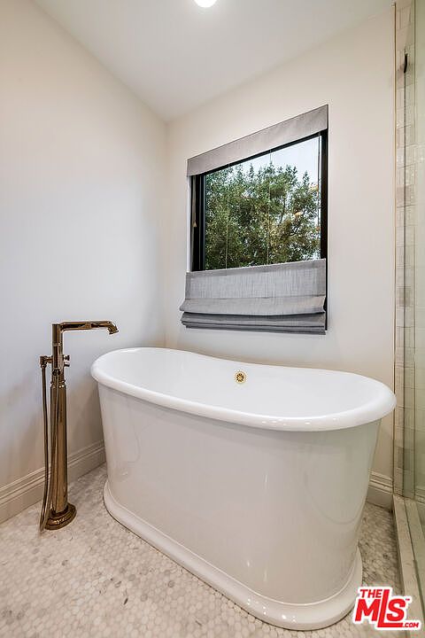 This is a bright and elegant primary bathroom featuring a freestanding white bathtub with a gold faucet. A large window with a gray roman shade provides natural light and a view of the trees outside. The floor is tiled with small, light-colored hexagonal tiles, adding a touch of classic charm.