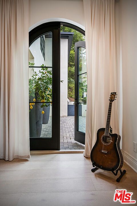 This interior shot showcases an elegant living space with a focus on natural light and a relaxed atmosphere. A dark wood acoustic guitar stands on a stand near an arched doorway with black trim, which leads to an outdoor patio. Light-colored curtains frame the doorway, adding softness to the room, and the light wood flooring contributes to the bright and airy feel.