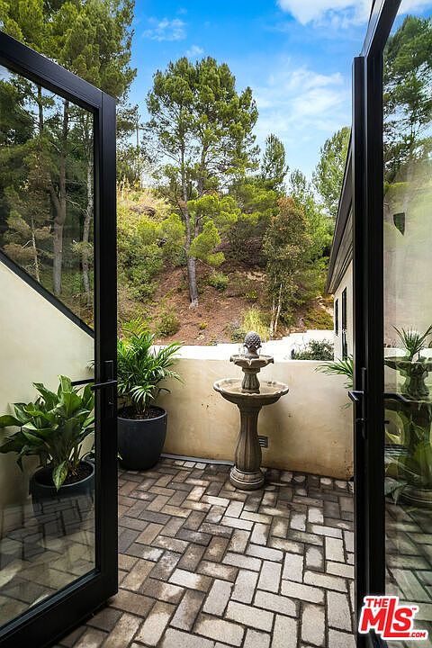This image showcases a charming patio area, accessible through black-framed glass doors, featuring a herringbone brick floor and a decorative stone fountain. Potted plants add greenery to the space, while the backdrop reveals a lush hillside with mature trees. The patio offers a private and serene outdoor living space.