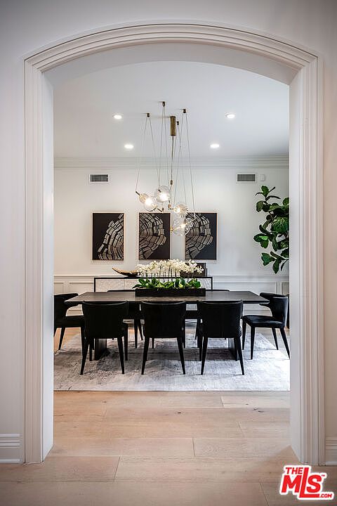 This image showcases a dining room viewed through an arched doorway, featuring a long, dark-colored dining table with black chairs. A modern chandelier hangs above the table, and artwork adorns the wall behind a sideboard. The room is well-lit and exudes a sophisticated, contemporary style.