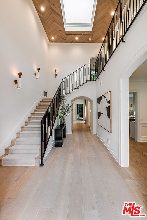This interior shot showcases a grand hallway with a staircase. The space features light wood flooring, white walls, and a black metal railing on the staircase. A large piece of modern art hangs on the wall, and sconces provide soft lighting, creating an elegant and inviting atmosphere.