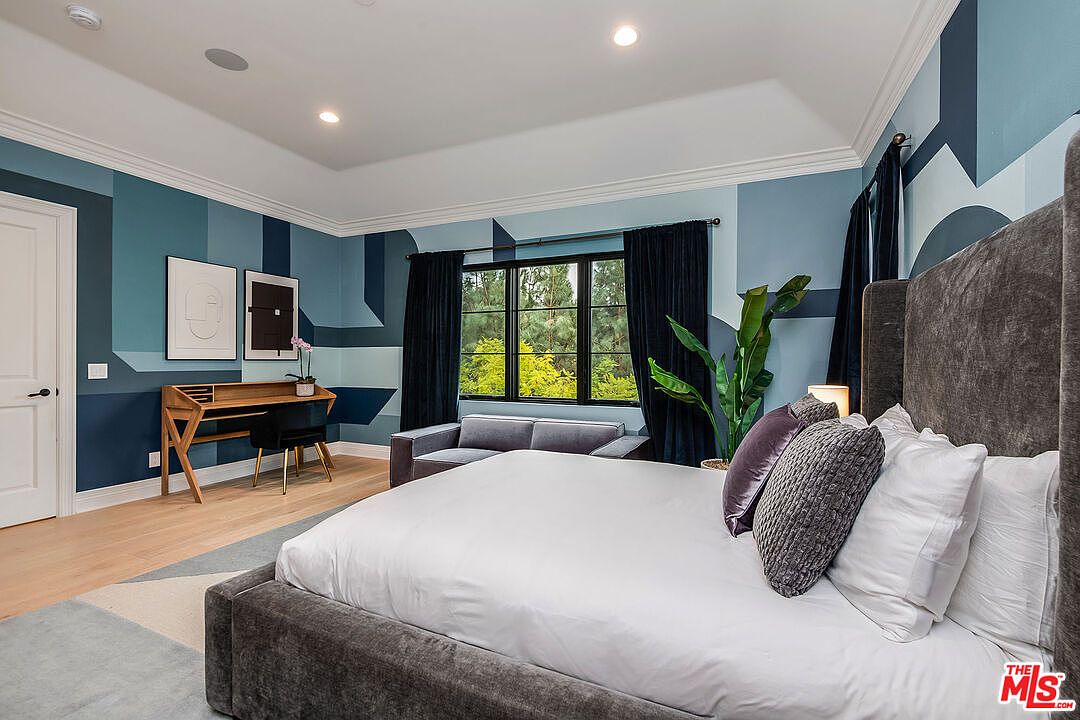This is a primary bedroom featuring a large bed with a gray upholstered frame and white linens. The walls are painted in a geometric pattern of blue hues, complemented by dark curtains. A desk and chair are positioned near a window, and a gray sofa sits in the background, creating a stylish and comfortable space.