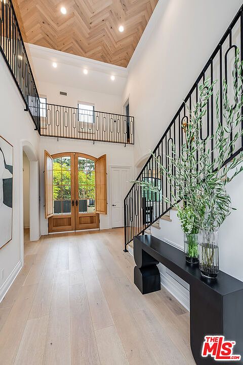 This is an interior shot of a grand hallway featuring light wood flooring and a high ceiling with a herringbone pattern. A staircase with a black railing leads to a second-floor balcony, also with a black railing. Double doors with shutters provide natural light and a view to the outside, while a sleek black console table with floral arrangements adds a modern touch.