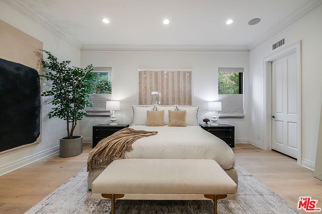 This is a well-lit primary bedroom featuring a large bed with neutral-toned bedding and decorative pillows. Two dark wood nightstands flank the bed, each with a lamp. A beige bench sits at the foot of the bed, and a large abstract painting and potted plant add visual interest. The room has hardwood floors and white walls, creating a serene and inviting atmosphere.