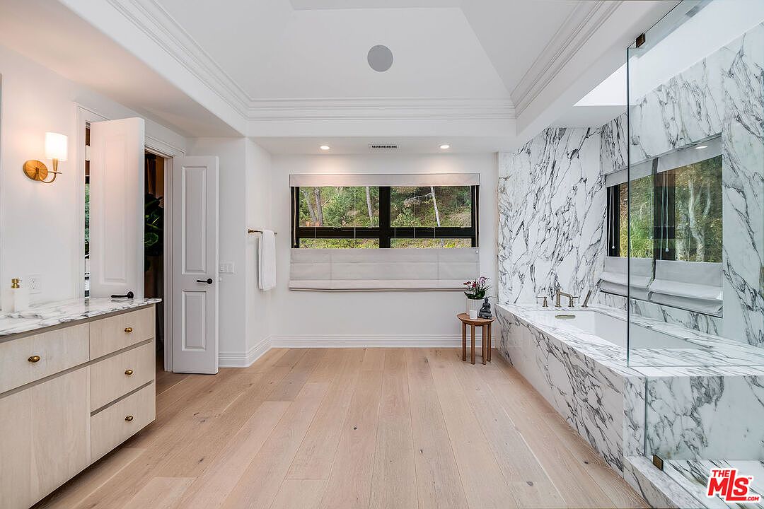 This is a luxurious primary bathroom featuring marble walls and a marble-clad bathtub. A large window provides natural light and a view of the outdoors. The bathroom also includes a vanity with a marble countertop and light wood cabinets, complemented by light hardwood flooring, creating a bright and elegant space.