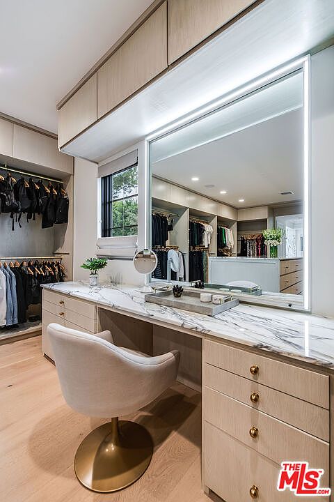This is an interior shot of a luxurious walk-in closet featuring a vanity area. The vanity has a marble countertop, a large mirror with integrated lighting, and a comfortable swivel chair. The closet also includes custom cabinetry and hanging racks for clothing, creating an organized and elegant space.