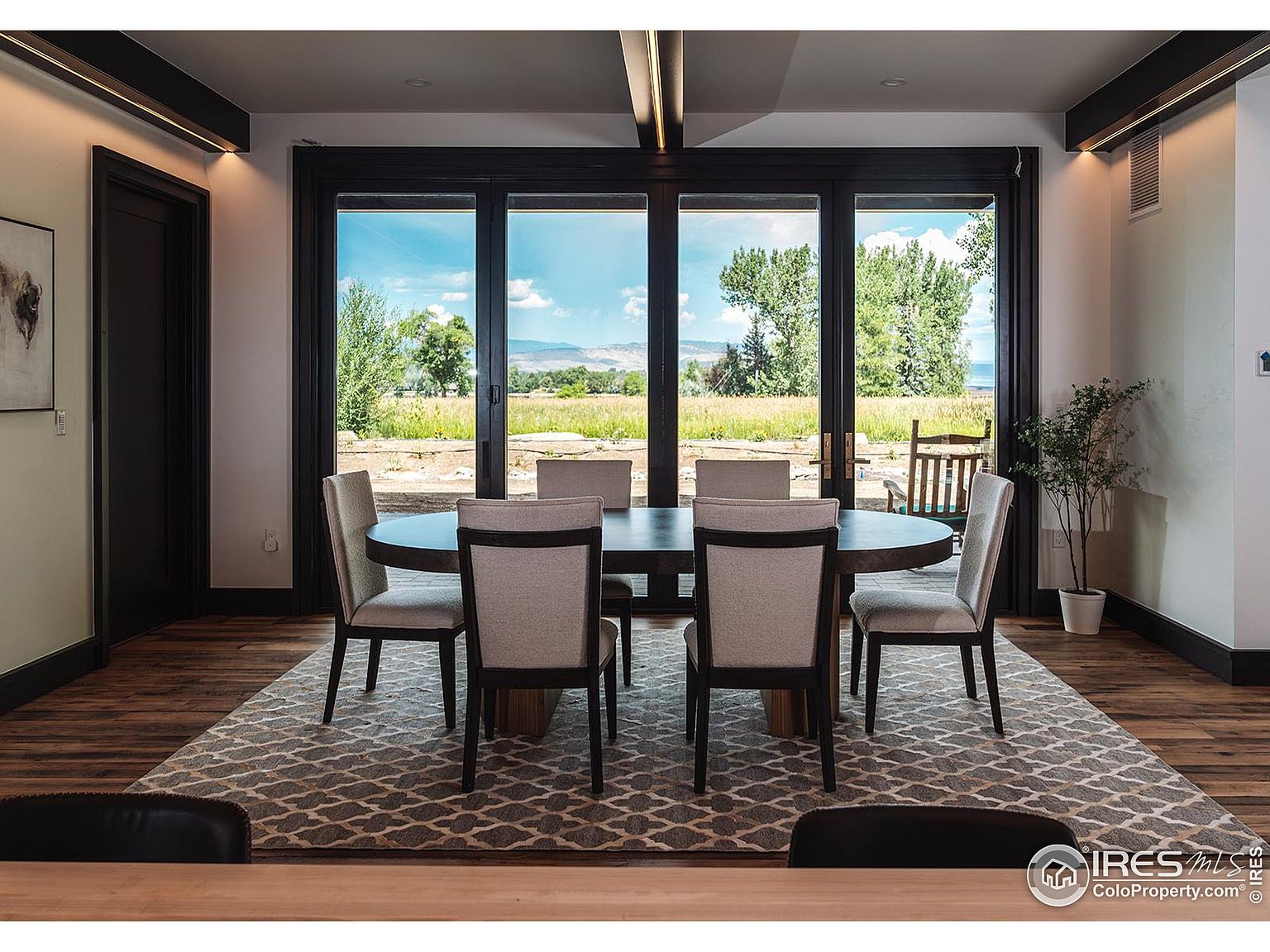 This is an interior shot of a dining room featuring a dark wood oval table surrounded by six upholstered chairs. A large area rug sits beneath the table, and a wall of sliding glass doors provides a view of a grassy field and distant mountains. The room is well-lit, creating a warm and inviting atmosphere.