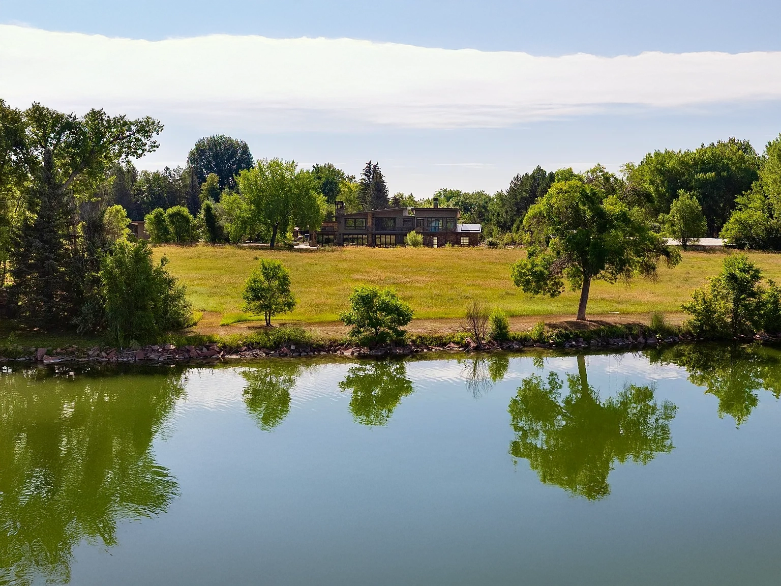 This aerial view showcases a single-family home situated on a vast, well-maintained lawn extending to the water. The modern architecture of the house is visible amidst the surrounding trees. The serene lake and the expansive green space emphasize the property's privacy and natural beauty, suggesting a tranquil and upscale living environment.