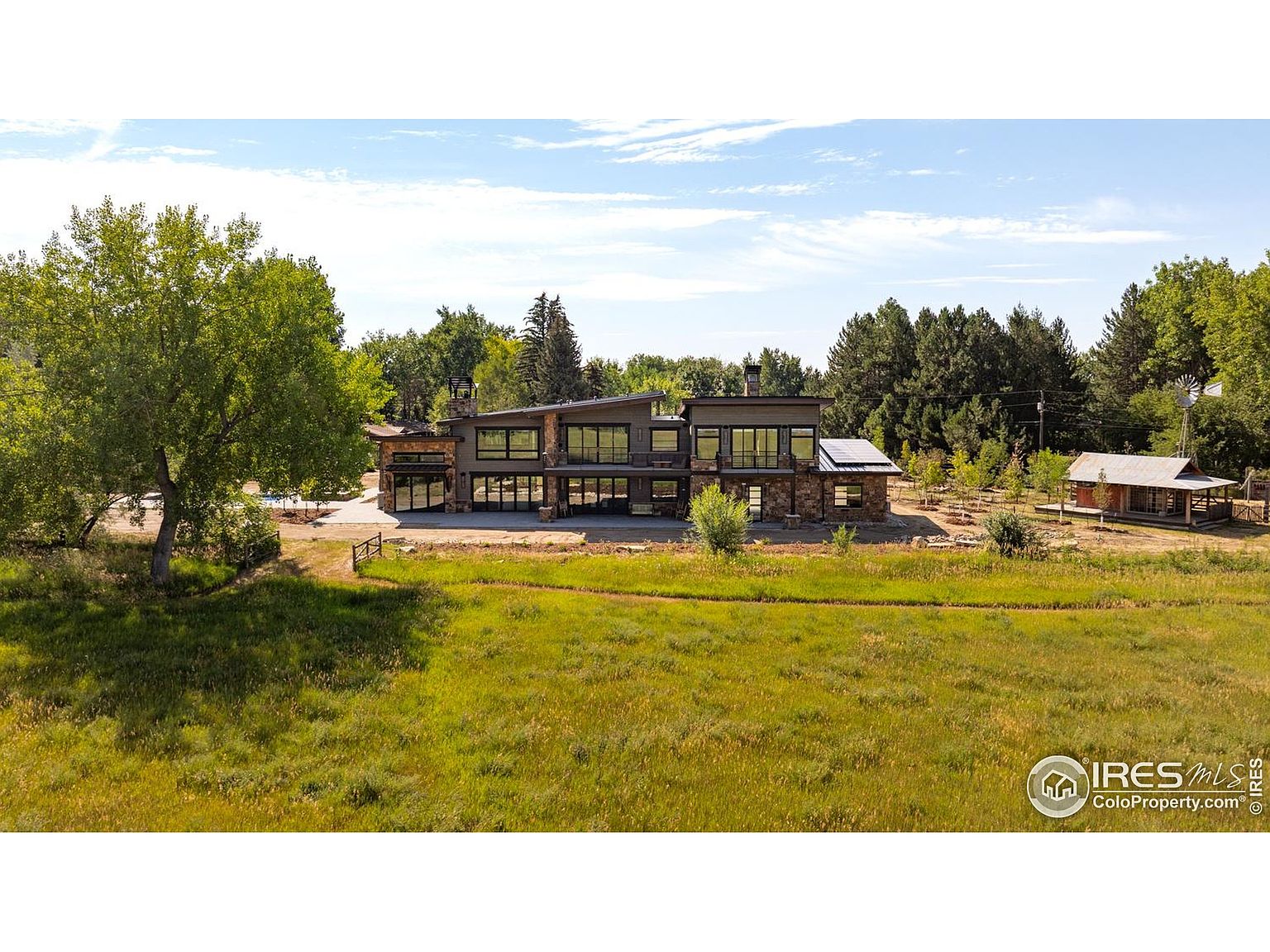 This aerial view showcases a modern, two-story home with a stone and dark gray exterior, surrounded by a lush green lawn and mature trees. The property includes a separate outbuilding, possibly a guest house or workshop, adding to the estate's appeal. The overall impression is one of privacy and luxury in a serene, natural setting.