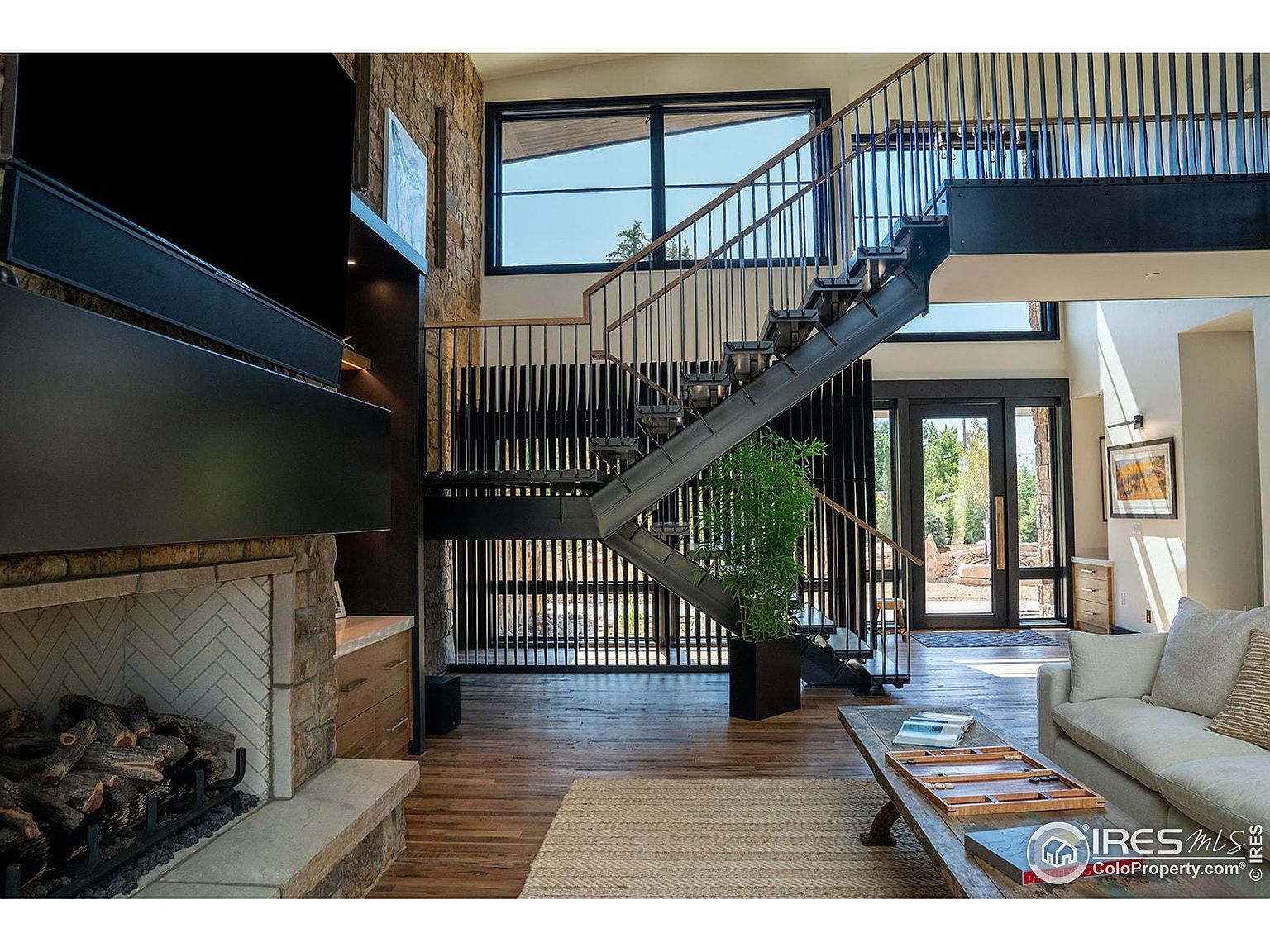 This is an interior shot of a living room featuring a modern staircase with metal railings, a stone fireplace with a herringbone pattern, and a comfortable-looking sofa. Large windows provide ample natural light, and the hardwood floors add warmth to the space. The overall impression is one of contemporary elegance and comfort.
