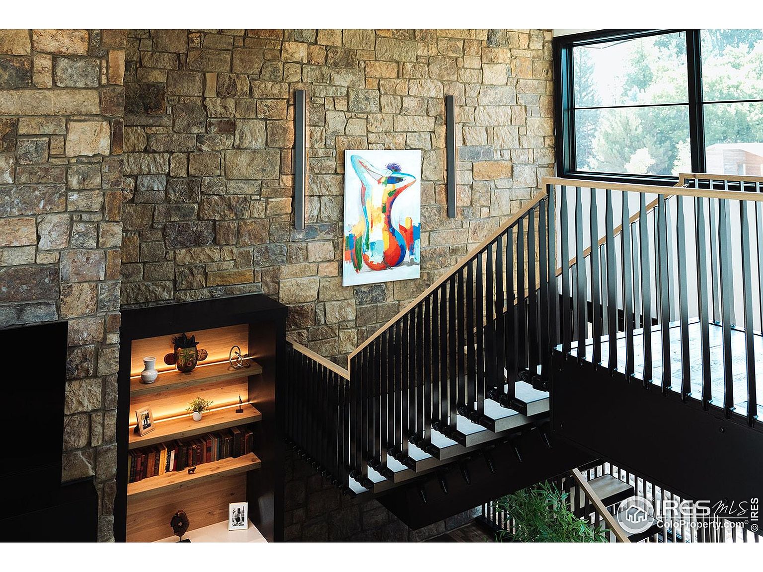 This interior shot showcases a modern hallway and staircase with a striking stone wall as a focal point. The staircase features dark railings and light-colored steps, creating a visually appealing contrast. An abstract painting and sleek light fixtures adorn the stone wall, adding an artistic touch to the space.