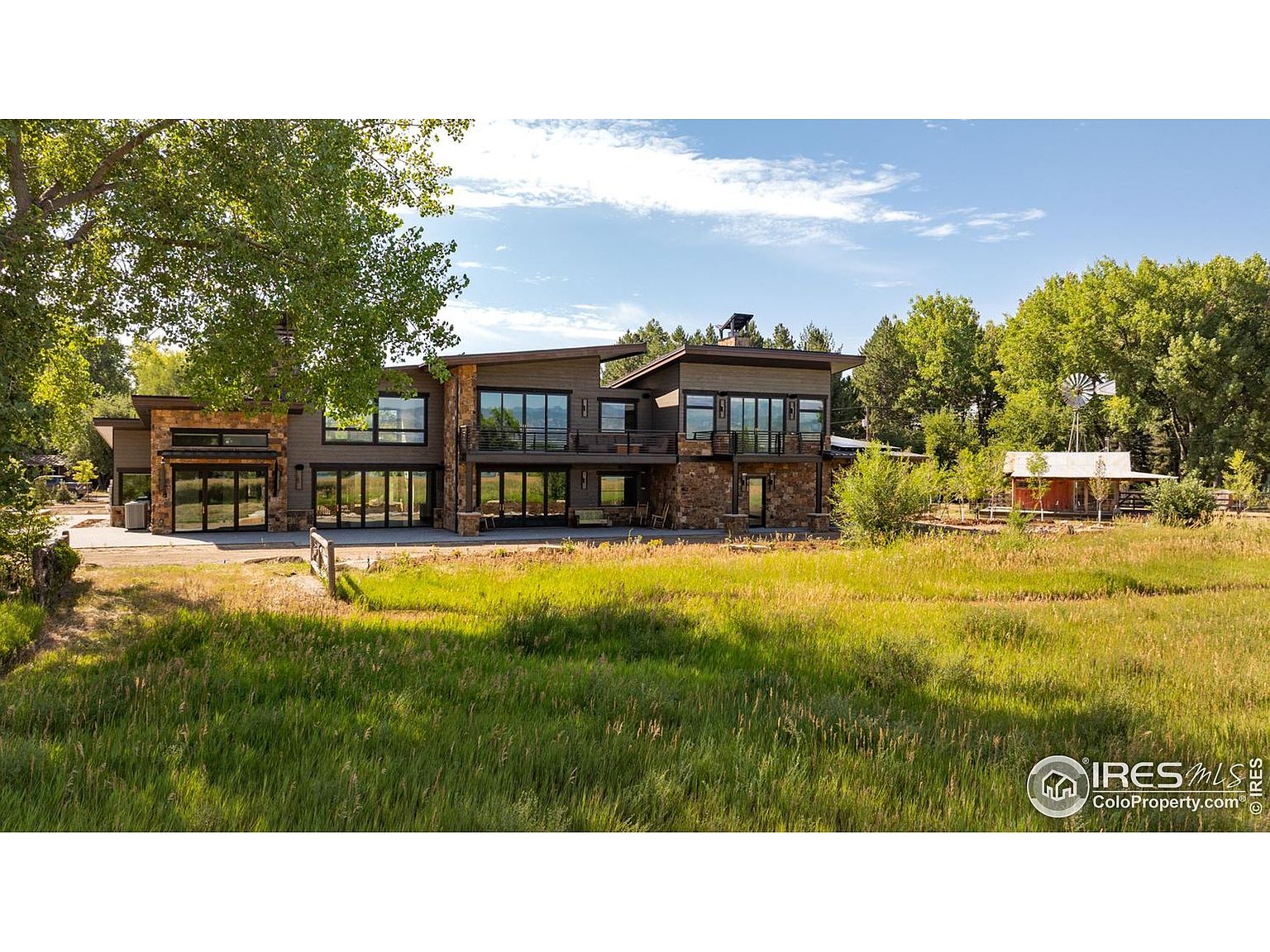 This image showcases the rear exterior of a modern home with a blend of natural and contemporary design elements. The house features large windows and balconies, stone accents, and a neutral color palette. The surrounding landscape includes a lush green lawn and mature trees, creating a serene and private setting.