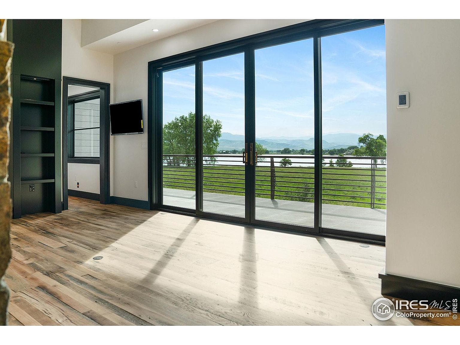 The image showcases a bright living room with hardwood floors and a large sliding glass door that opens to a balcony with a view of a lake and mountains. A built-in bookshelf and a wall-mounted television add to the room's functionality. The room exudes a modern and airy feel, emphasizing the connection to the outdoor scenery.