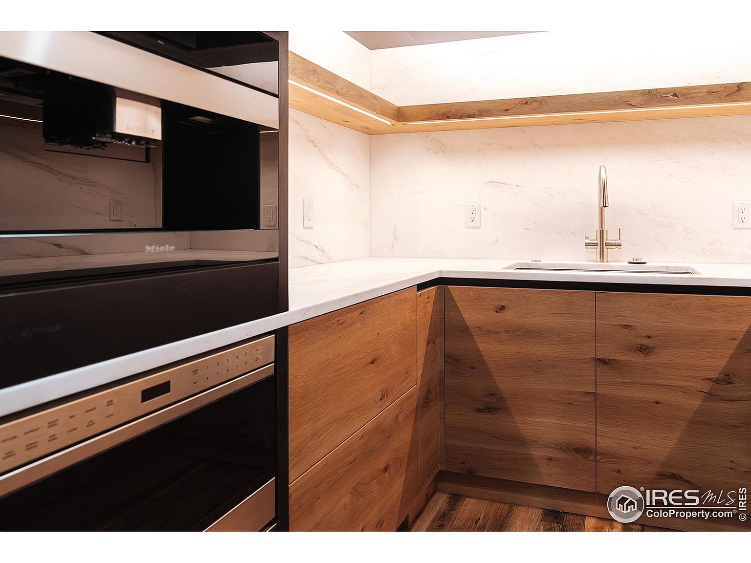 This image showcases a modern kitchen featuring sleek, wood-grain cabinetry and a light-colored countertop. A Miele oven and built-in coffee machine are visible, adding a touch of luxury. The under-cabinet lighting and marble backsplash contribute to a clean and sophisticated aesthetic.