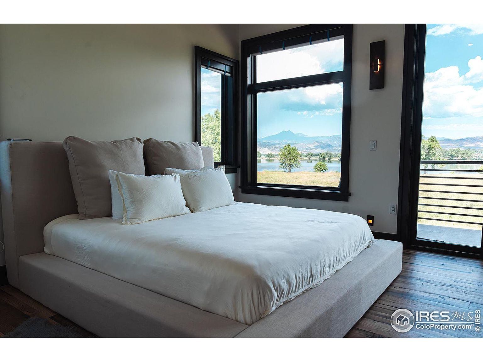 This is a primary bedroom featuring a large platform bed with neutral-toned linens and pillows. The room has large windows offering a scenic view of a lake and mountains, enhancing the sense of tranquility. Dark window frames provide a modern contrast to the light walls and bedding, and the hardwood floors add warmth.