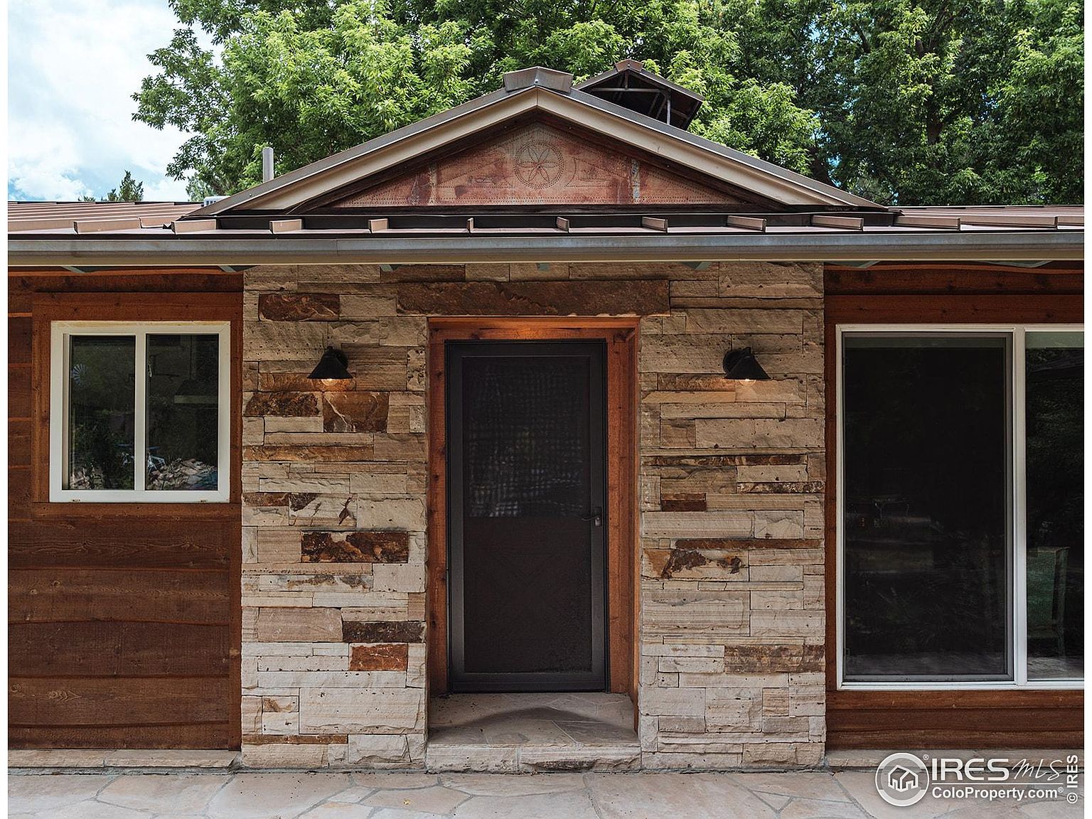 This image showcases the entryway of a home, featuring a stone facade, a dark-colored front door with a screen, and symmetrical sconce lighting. The entrance is framed by wood trim, and there are windows on either side, creating a welcoming and well-lit approach. The overall style is rustic and inviting.