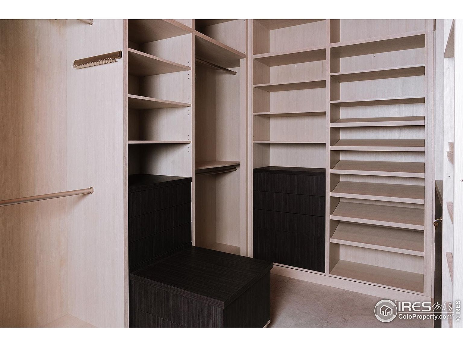 This is an interior shot of a well-organized walk-in closet. The closet features a combination of open shelving, drawers, and hanging rods, providing ample storage space. The light-colored wood finish gives the closet a clean and modern look, while the dark drawers add a touch of contrast.