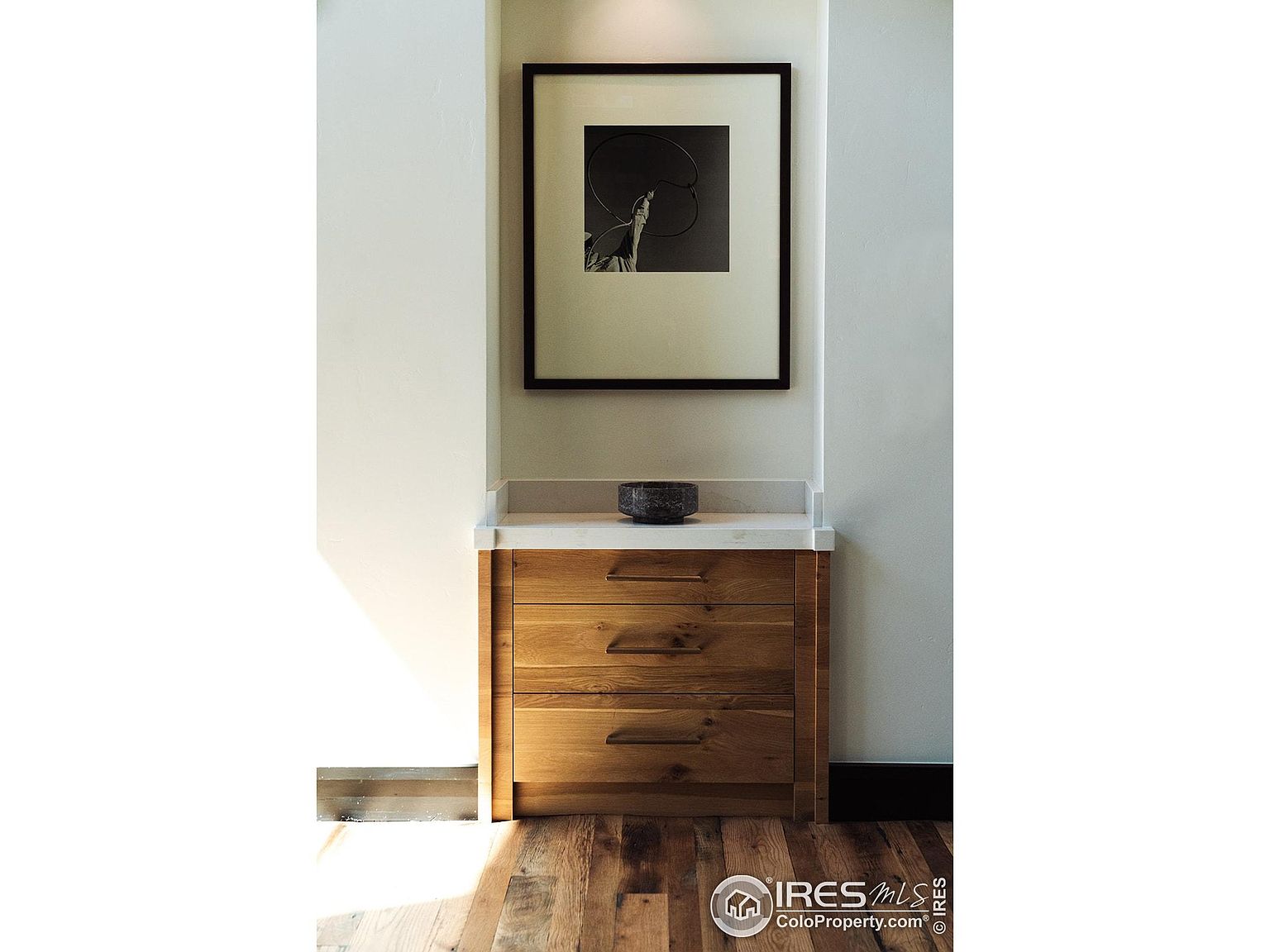 This interior shot showcases a built-in wooden cabinet with three drawers and a light countertop, set within a recessed wall niche. Above the cabinet hangs a framed black and white artwork, adding a touch of sophistication. The hardwood flooring and the play of light and shadow create a warm and inviting atmosphere.
