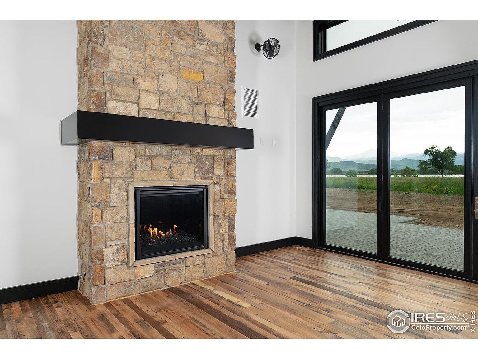 This interior shot showcases a living room with a prominent stone fireplace and a dark wood mantel. The room features hardwood flooring and a large sliding glass door that provides ample natural light and a view of the outdoors. The overall aesthetic is modern with rustic elements, creating a warm and inviting atmosphere.