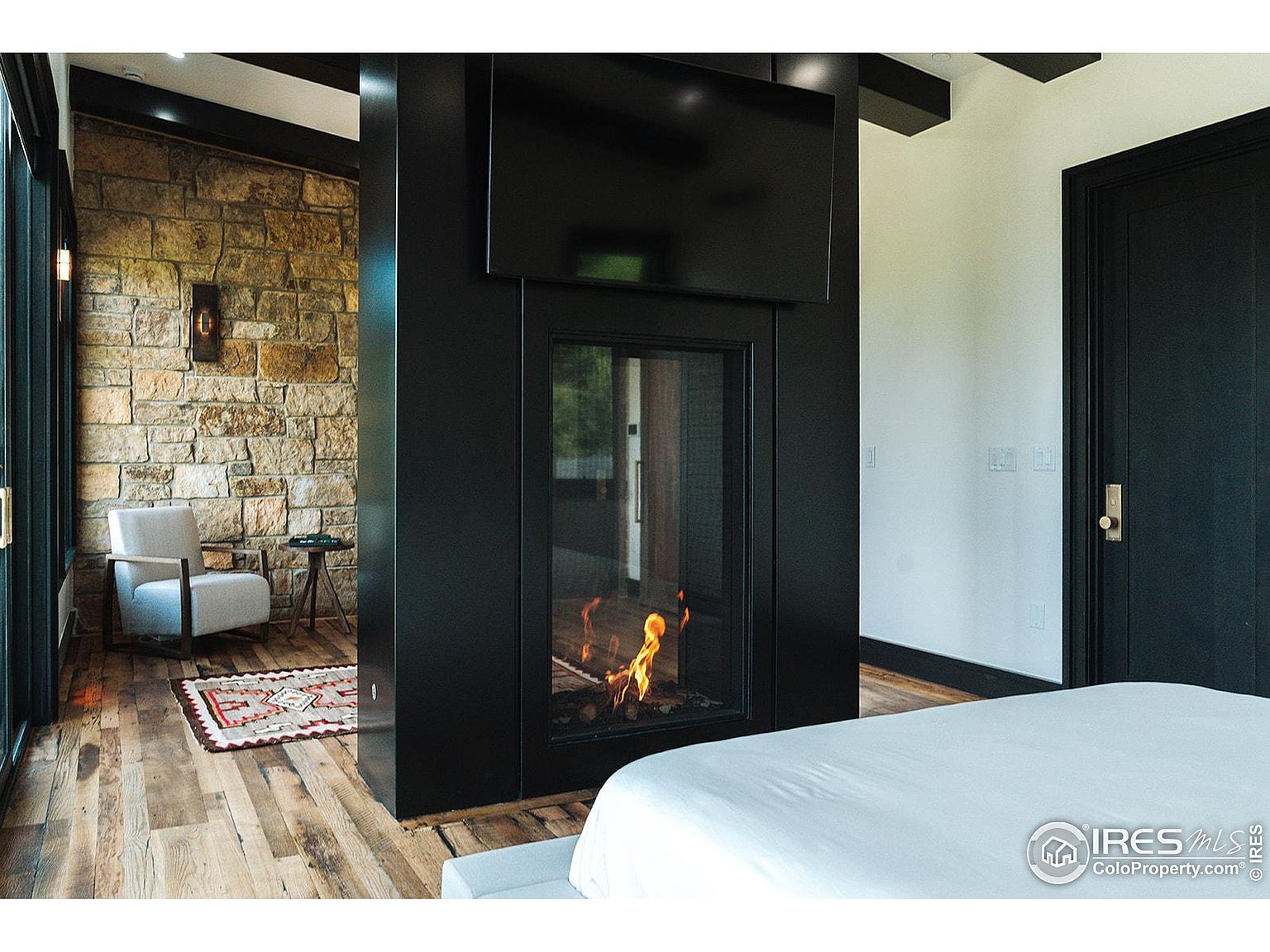 This is a primary bedroom featuring a modern fireplace with a mounted television above it. The room has a stone accent wall, hardwood floors, and a black door. The bed is neatly made with white linens, and the overall style is contemporary and luxurious.