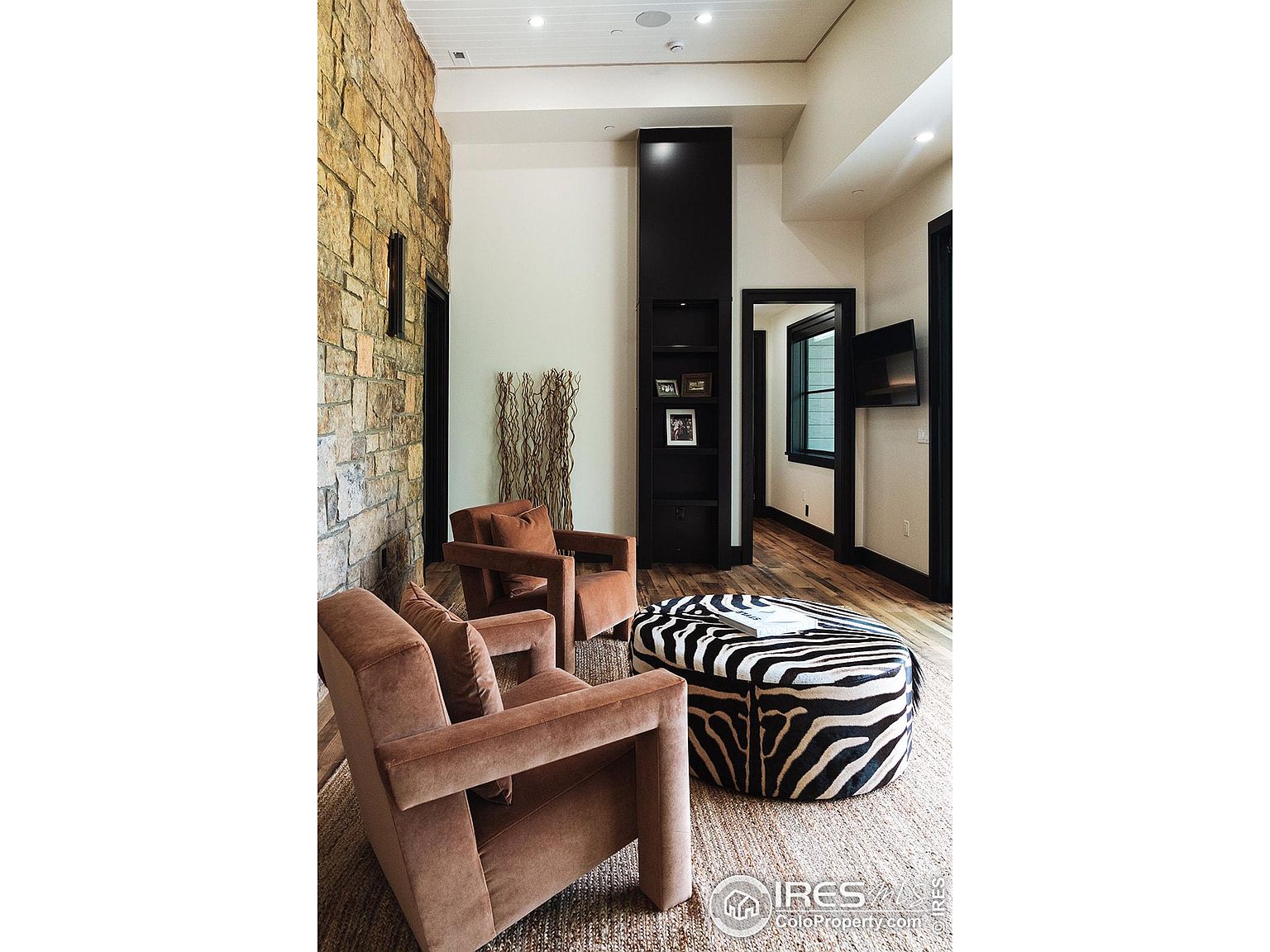 This interior shot showcases a stylish living room with a blend of textures and modern design. The room features two plush armchairs, a zebra-print ottoman, and a striking stone accent wall. A tall, dark shelving unit adds vertical interest, while the hardwood flooring and neutral rug create a warm and inviting atmosphere.