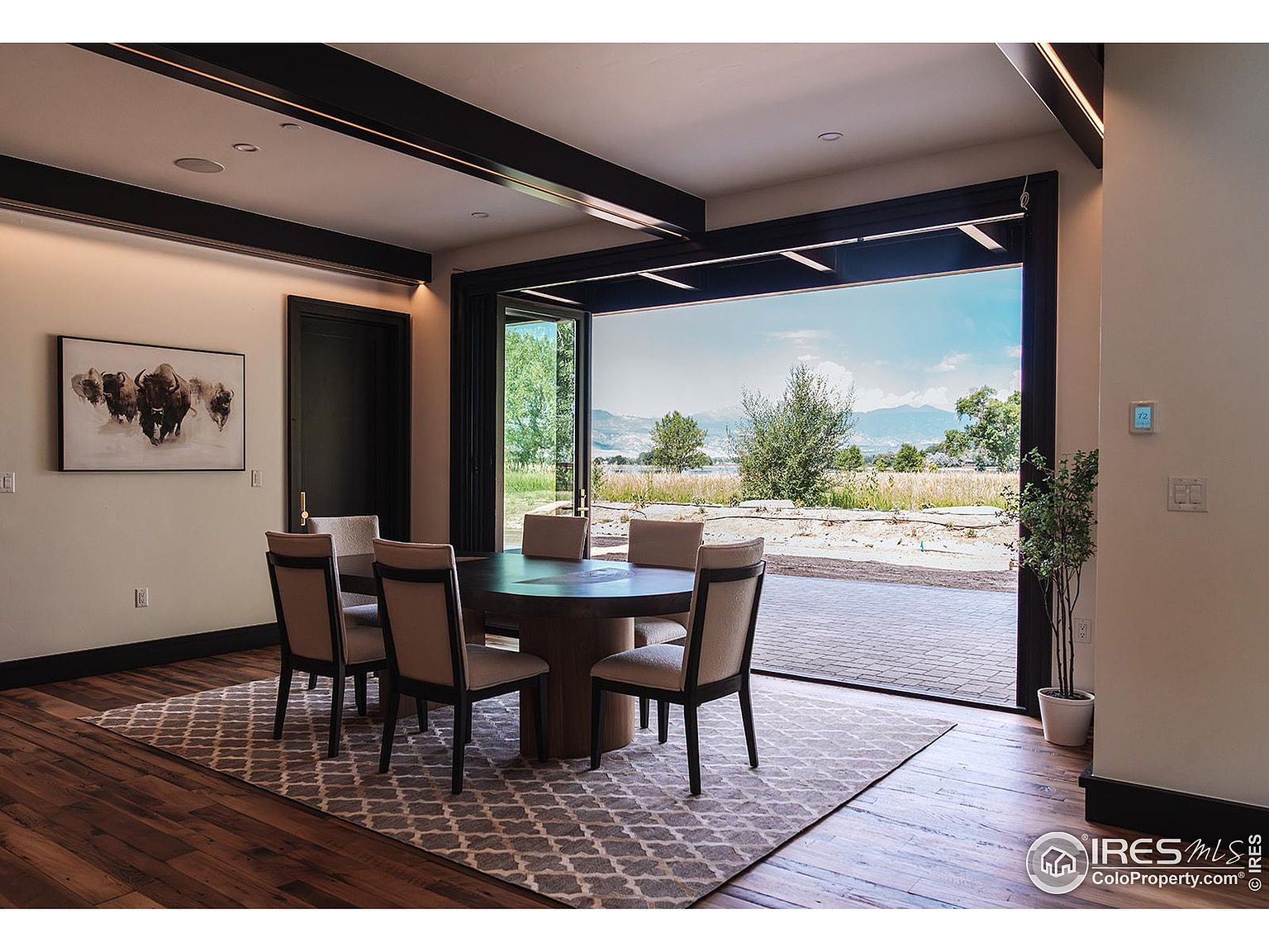 This is an interior shot of a dining room featuring a round wooden table with six chairs, set on a patterned rug over hardwood floors. A large, multi-panel glass door opens to an outdoor view of a grassy landscape with trees and distant mountains, creating a seamless indoor-outdoor living experience. The room is well-lit, with dark wood beams on the ceiling adding a touch of architectural detail.