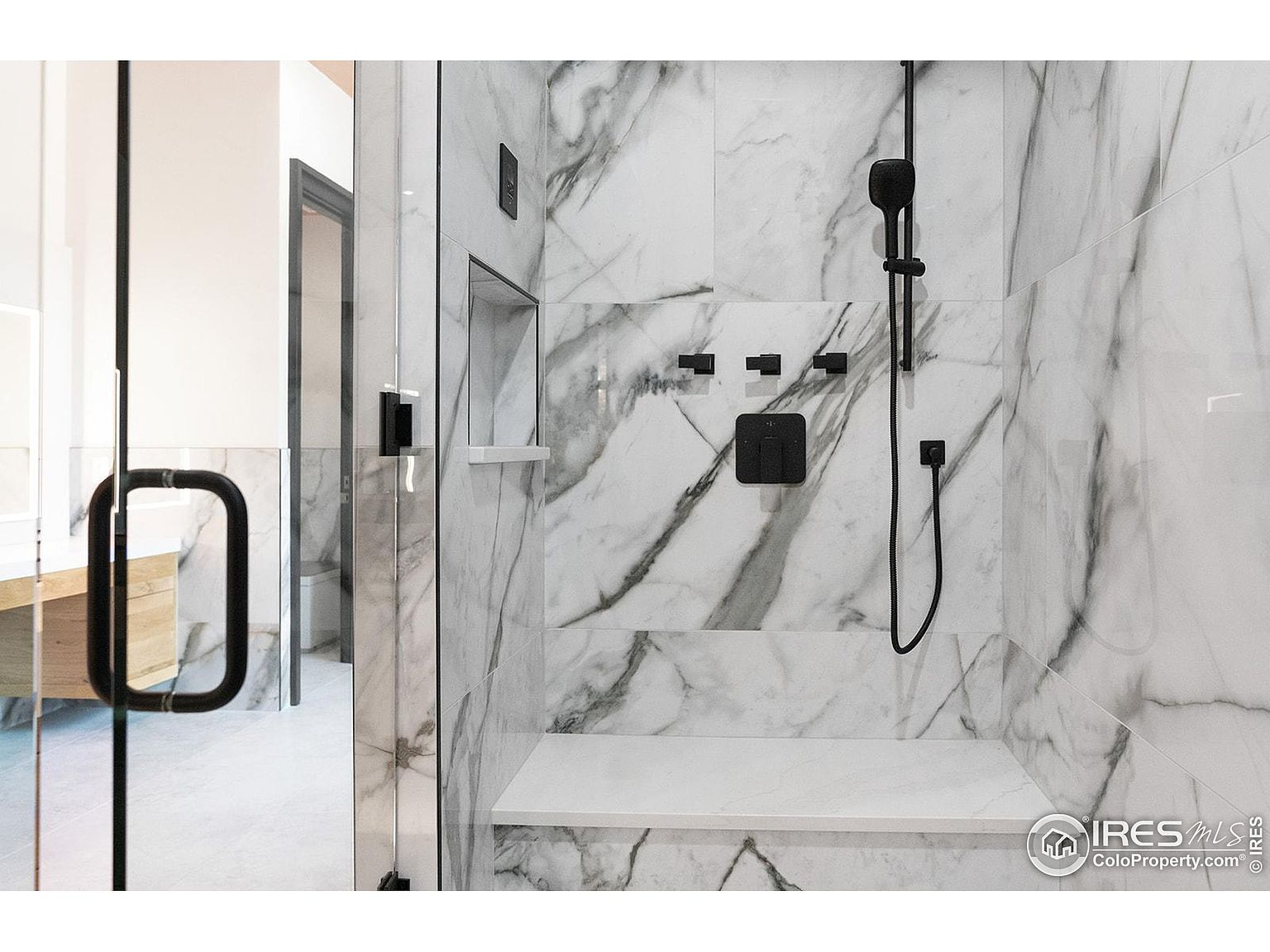 This image showcases a luxurious primary bathroom shower. The shower features marble-patterned tile walls, a built-in bench, and sleek black fixtures, including a shower head and controls. A glass door encloses the shower, providing a clear view of the bathroom beyond, which includes a vanity and toilet.