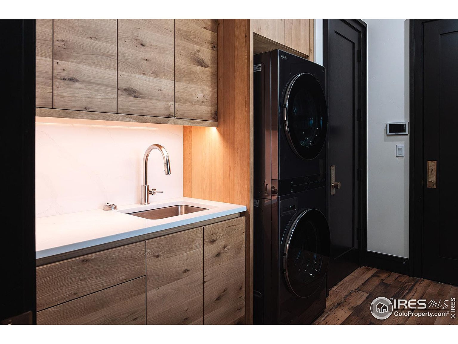 This is a well-appointed laundry room featuring modern wood cabinetry, a white countertop with a stainless steel sink and faucet, and a stacked black washer and dryer unit. A black door with brass hardware adds a touch of elegance, while the wood flooring complements the overall design. The room appears clean, organized, and functional, making it an attractive feature for potential buyers.