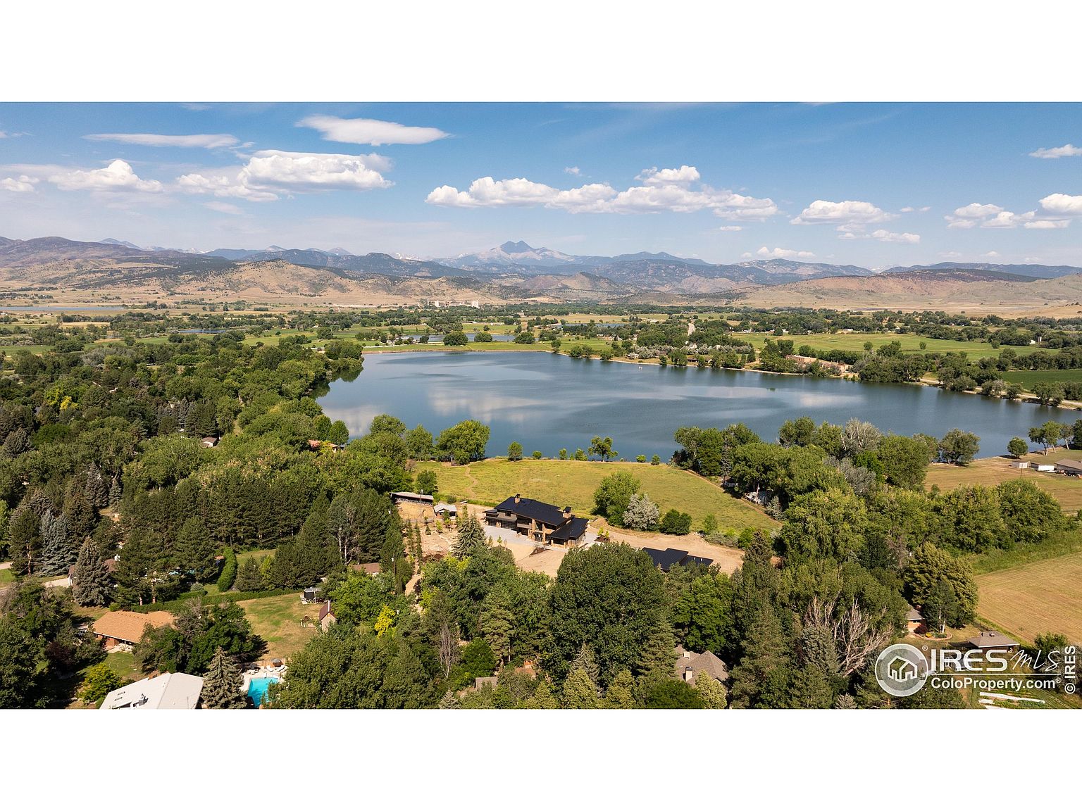 This aerial shot showcases a stunning property nestled beside a serene lake, surrounded by lush greenery and mature trees. The house features a dark roof and appears to have multiple structures, blending harmoniously with the natural landscape. In the background, rolling hills and distant mountains add depth and grandeur to the scene, creating a picturesque and desirable setting.