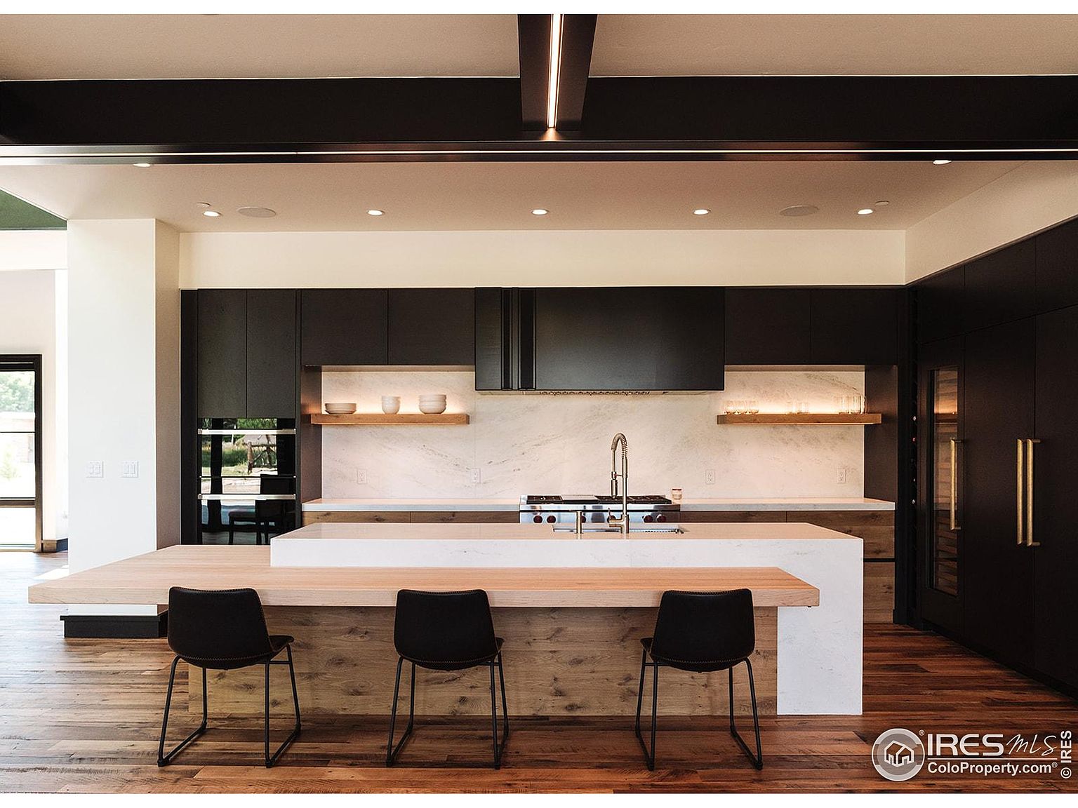 This is a modern kitchen featuring dark cabinetry, a light wood island with a white countertop, and stainless steel appliances. The kitchen has a clean and minimalist design, with open shelving and a marble backsplash. The perspective is from the front, showcasing the island and the layout of the kitchen.