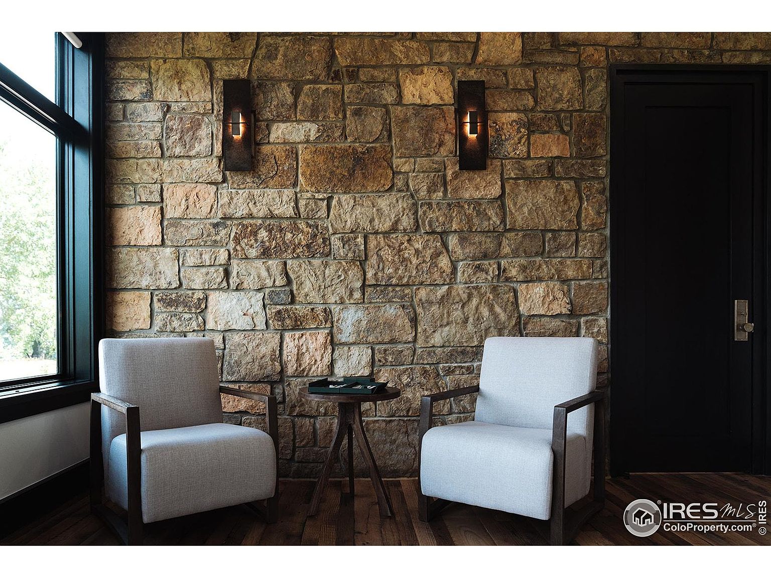 This interior shot showcases a cozy living room area with two armchairs flanking a small side table, set against a textured stone wall. Two sconces provide warm lighting, and a dark door adds a touch of contrast. The room exudes a rustic yet modern ambiance, perfect for a relaxing space.