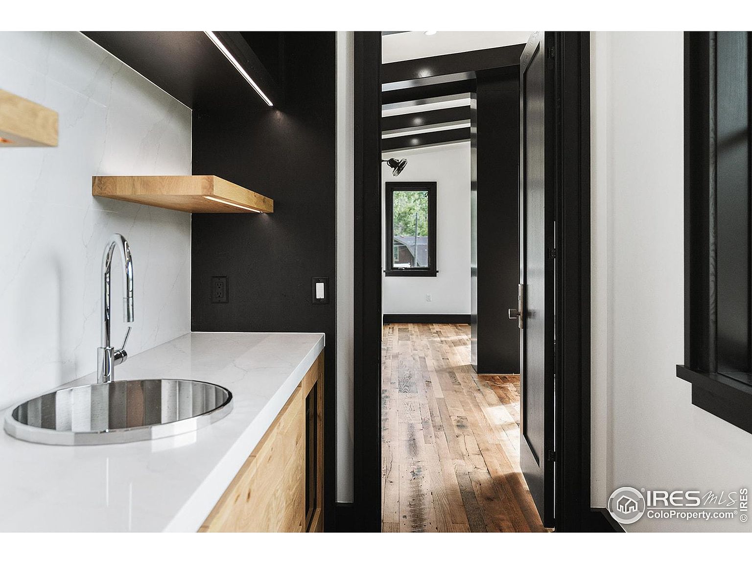 This image showcases a modern bathroom with a sleek design. The vanity features a white countertop, a stainless steel sink, and light wood cabinetry. Black accents are present on the walls and door frames, creating a striking contrast with the white walls and marble backsplash. The view through the doorway reveals hardwood flooring and a glimpse into another room, adding depth to the image.
