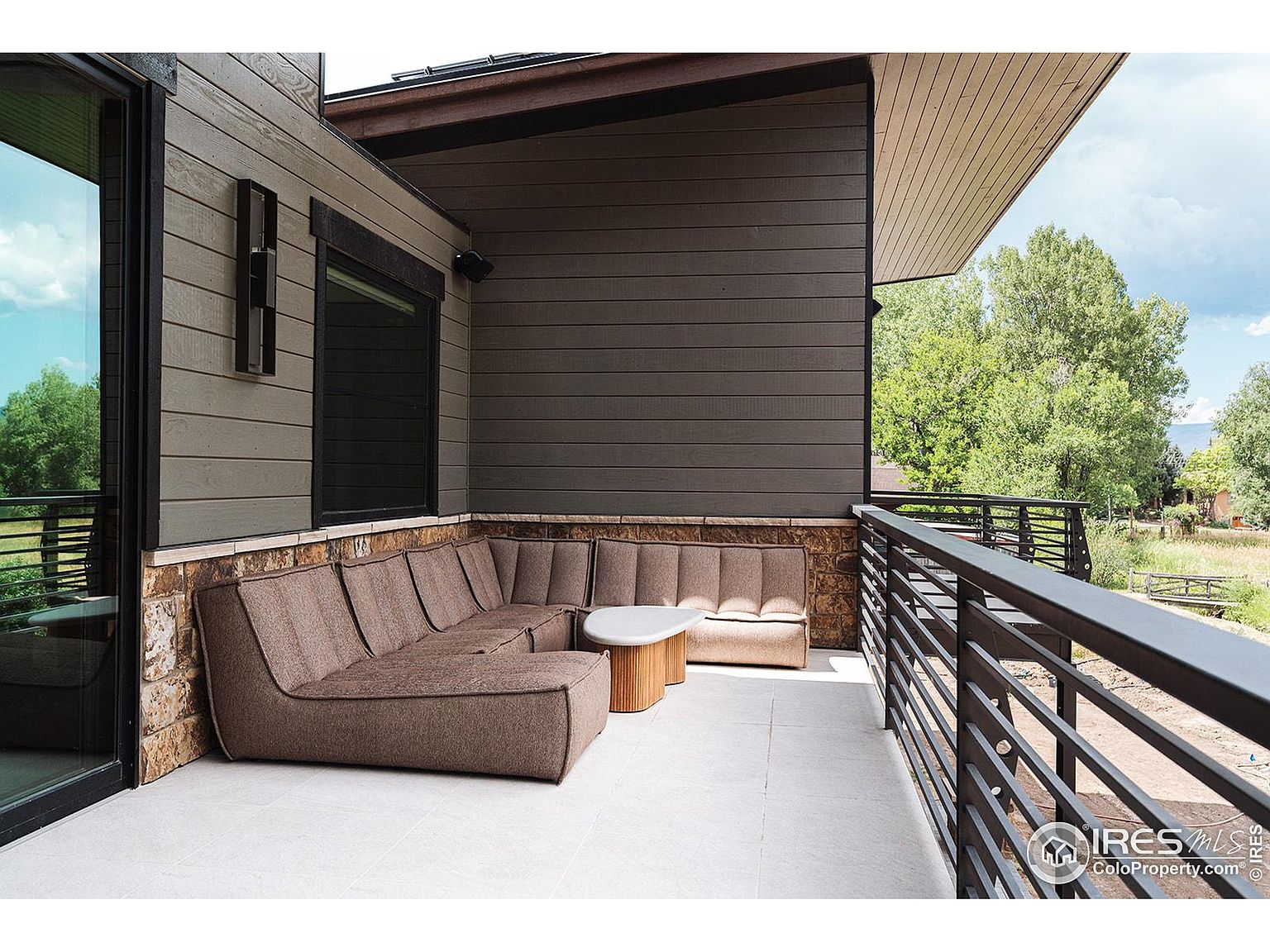This image showcases a stylish outdoor patio or balcony area. A modern, sectional sofa provides ample seating, complemented by a small, round coffee table. The space is enclosed by a sleek, horizontal railing, offering views of the surrounding greenery and landscape, creating an inviting and relaxing outdoor living space.
