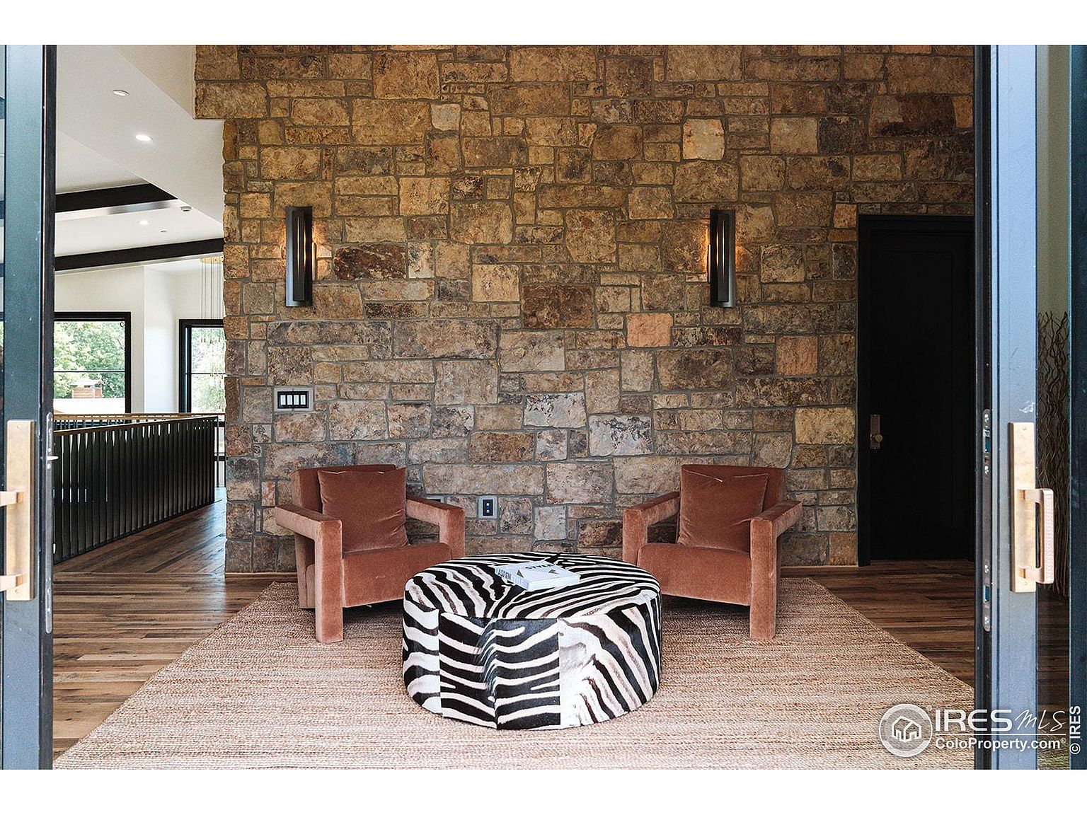 This interior shot showcases a stylish living room area with a striking stone wall, flanked by modern sconces. Two armchairs face a zebra-print ottoman, all set on a neutral rug, creating a cozy yet sophisticated seating arrangement. The open doorway provides a glimpse into another area of the home, enhancing the sense of space and flow.
